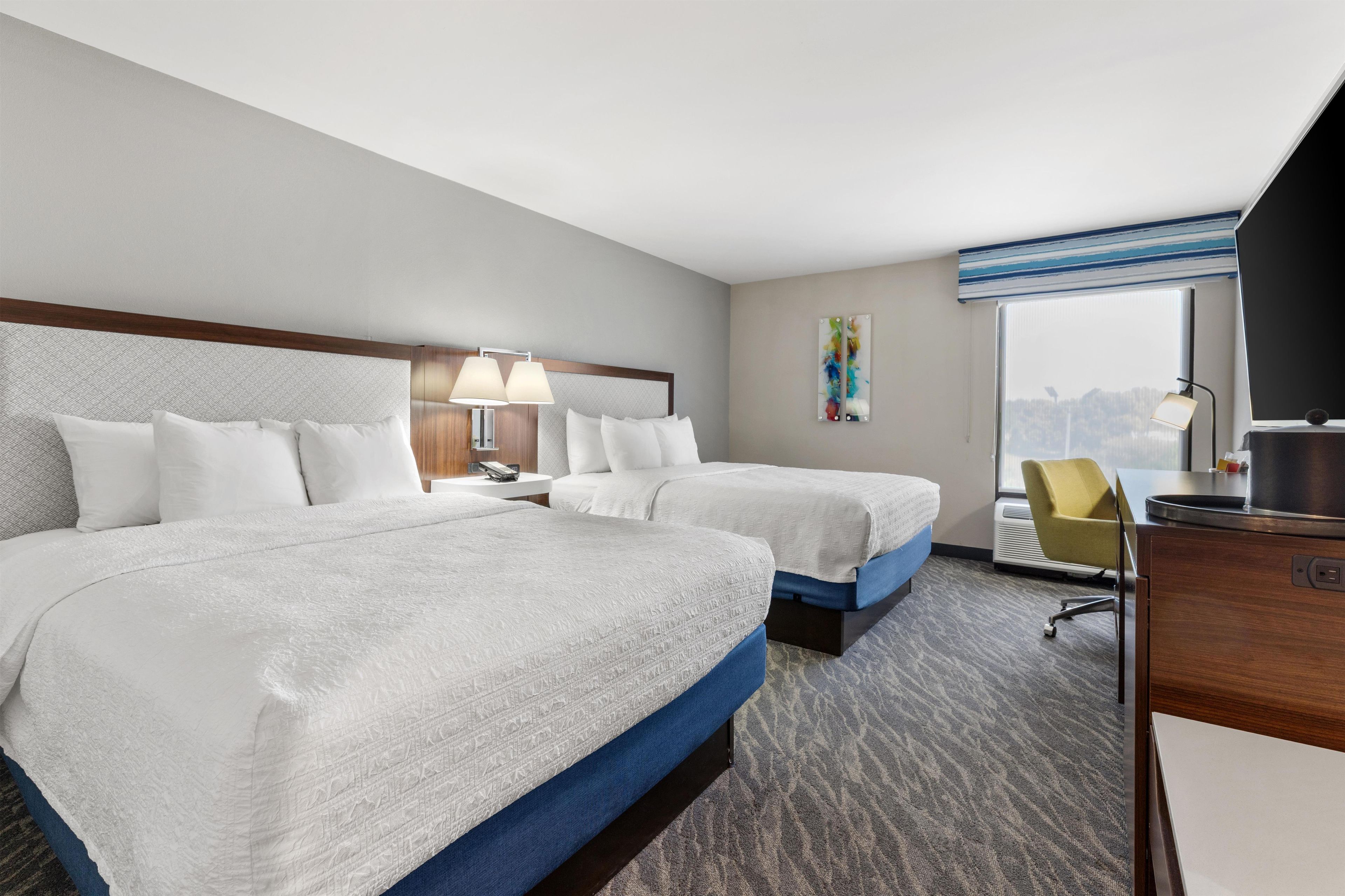 A hotel room with two beds, a desk, a chair, a window with a view, and a painting on the wall.