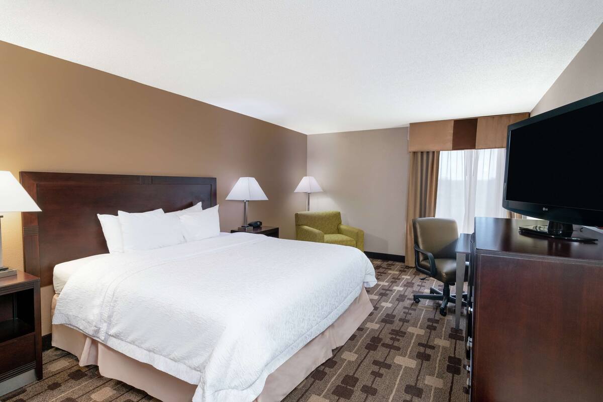 A hotel room with a large bed, two bedside lamps, a desk with a television, a yellow armchair, and a carpeted floor.