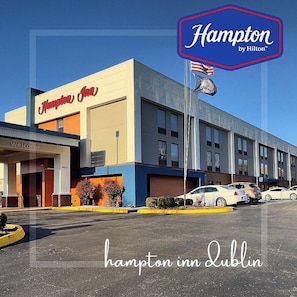 A Hampton Inn by Hilton building with a 'Welcome' sign, a parking lot, and several cars parked in front. - Hampton Inn Dublin (Dublin)
