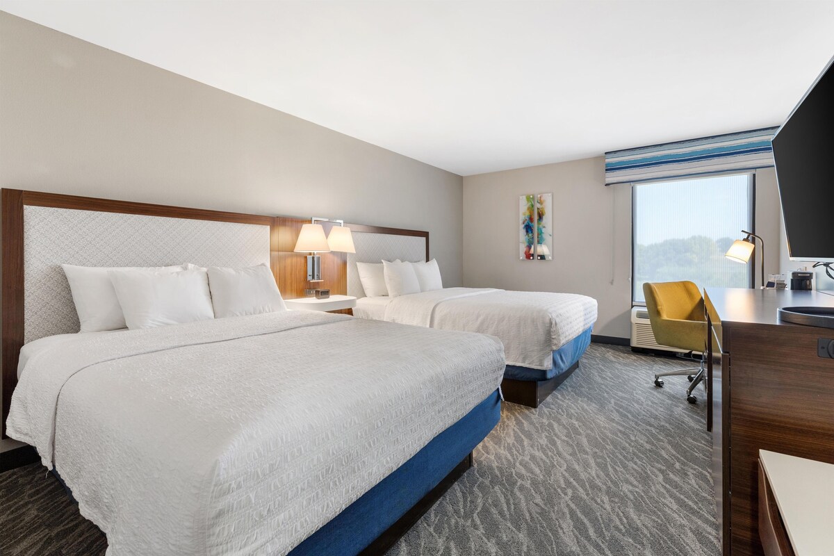 Habitación de hotel con dos camas, un escritorio, una silla y un ventanal grande.