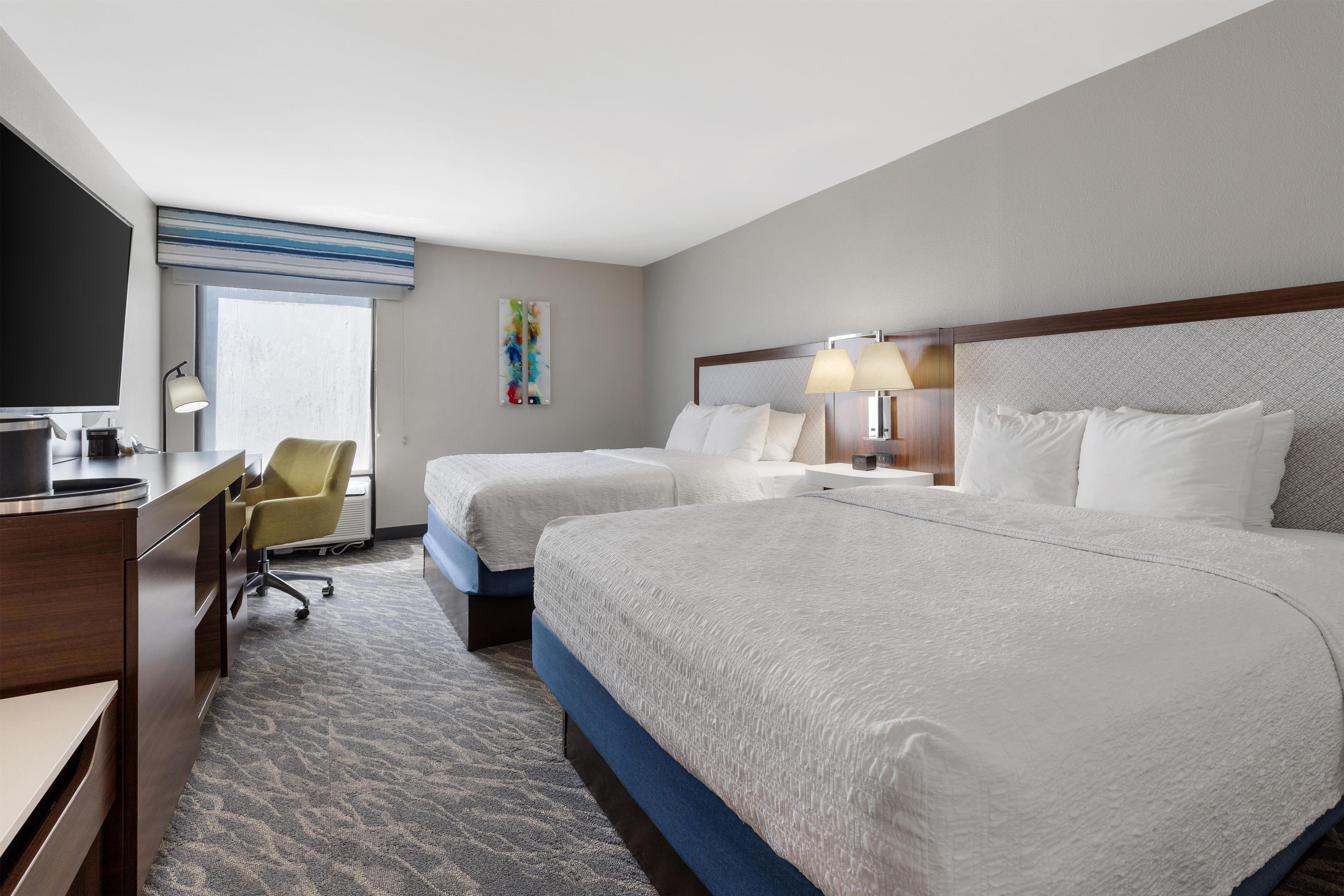 A hotel room with two beds, a desk, a chair, a television, and a window.