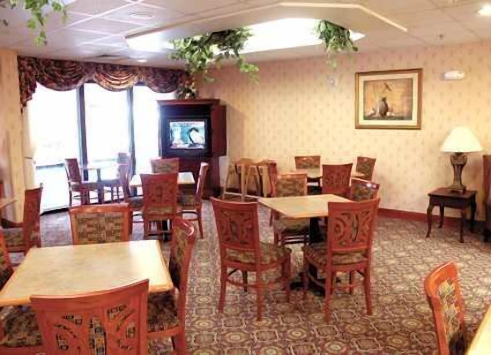 A hotel dining area with wooden tables and chairs, a television on the wall, and a patterned carpet.
