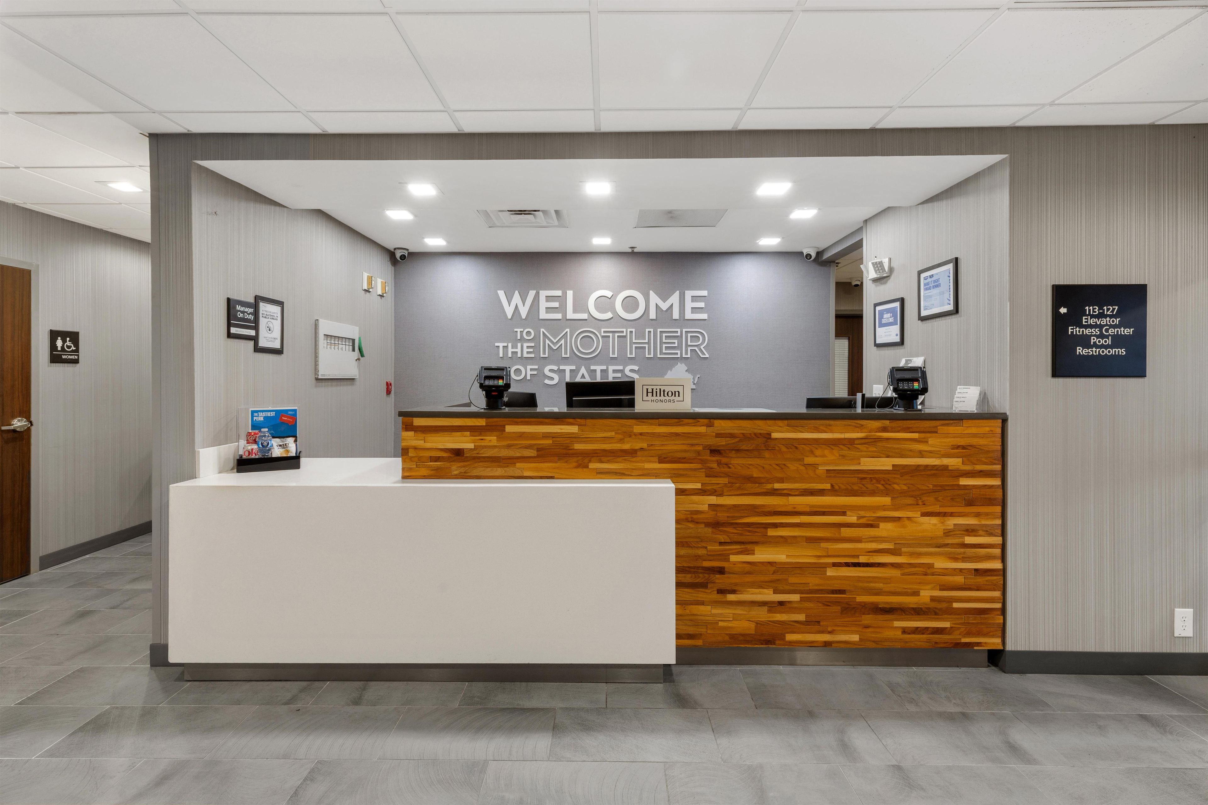 A reception desk with a 'WELCOME TO THE MOTHER OF STATES' sign, a 'Hilton Honors' badge, and directional signs for elevators and restrooms.