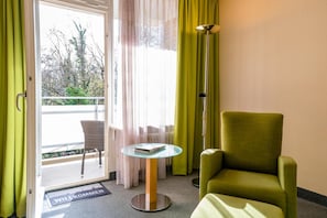 A room with a green armchair, a glass-top table with a book, a floor lamp, and a balcony door.