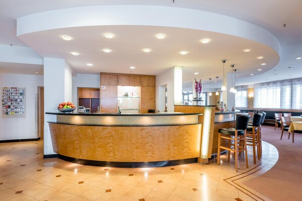 A modern hotel lobby with a curved reception desk, a kitchen area, and a seating area with chairs and tables.