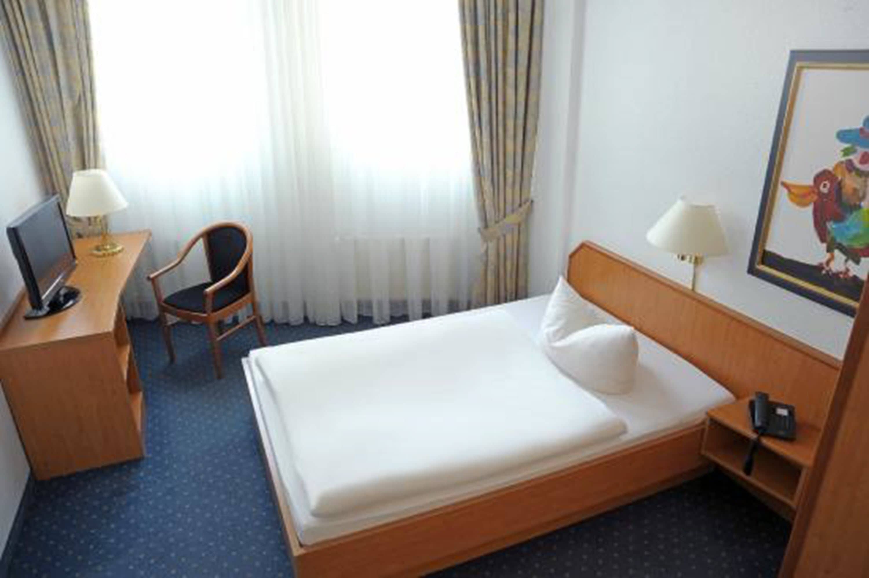Habitación de hotel con una cama individual, un escritorio con silla, una lámpara y un cuadro en la pared.