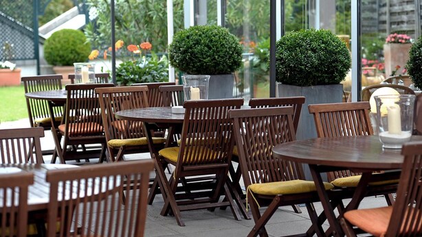 An outdoor patio area with round wooden tables and chairs, potted plants, and a glass railing. - Hotel Ludwig van Beethoven (Berlin)