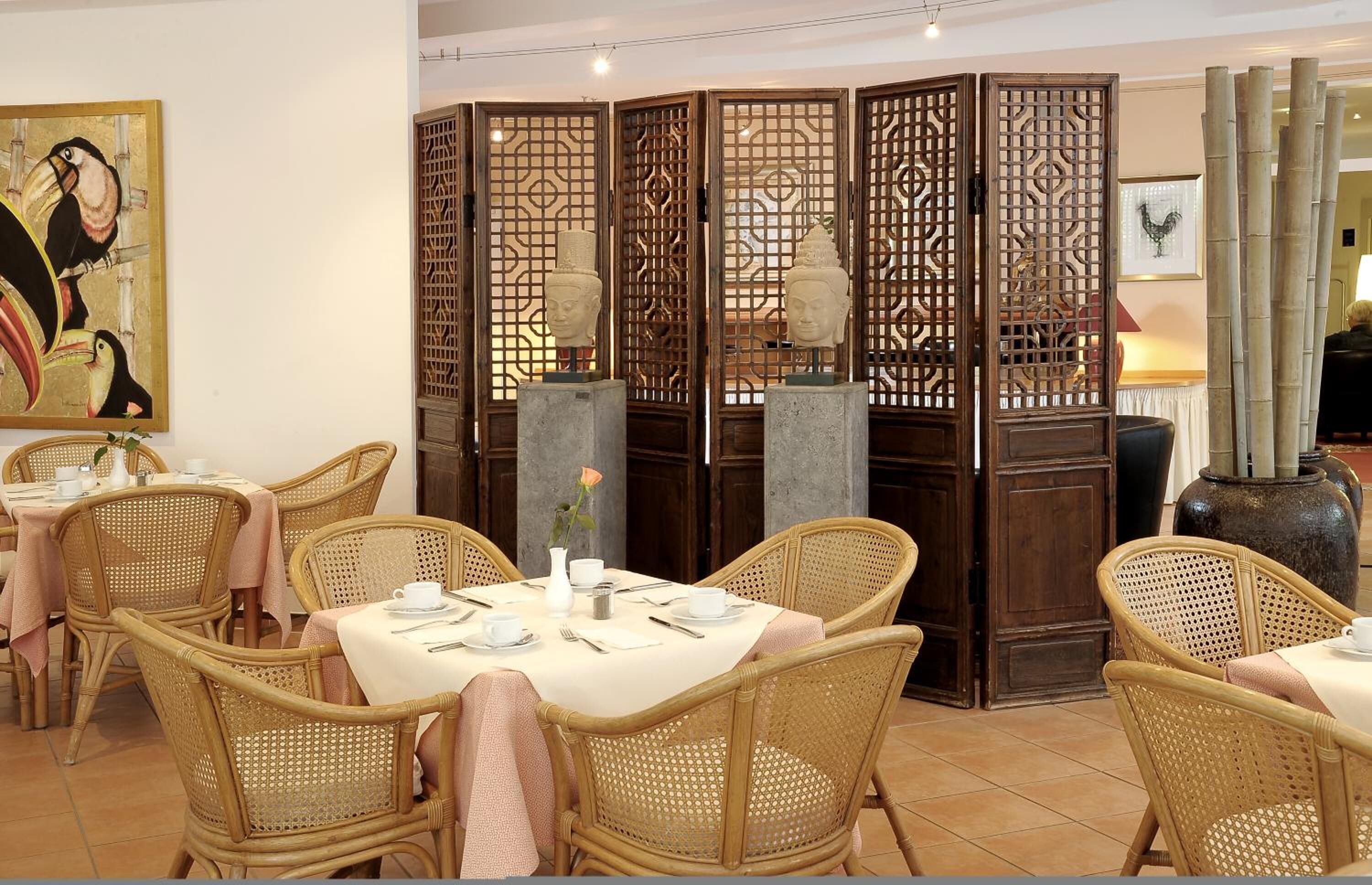 Ein Essbereich mit Korbstühlen und Tischen, einem dekorativen Raumteiler mit Buddha-Statuen und einer großen Vase.