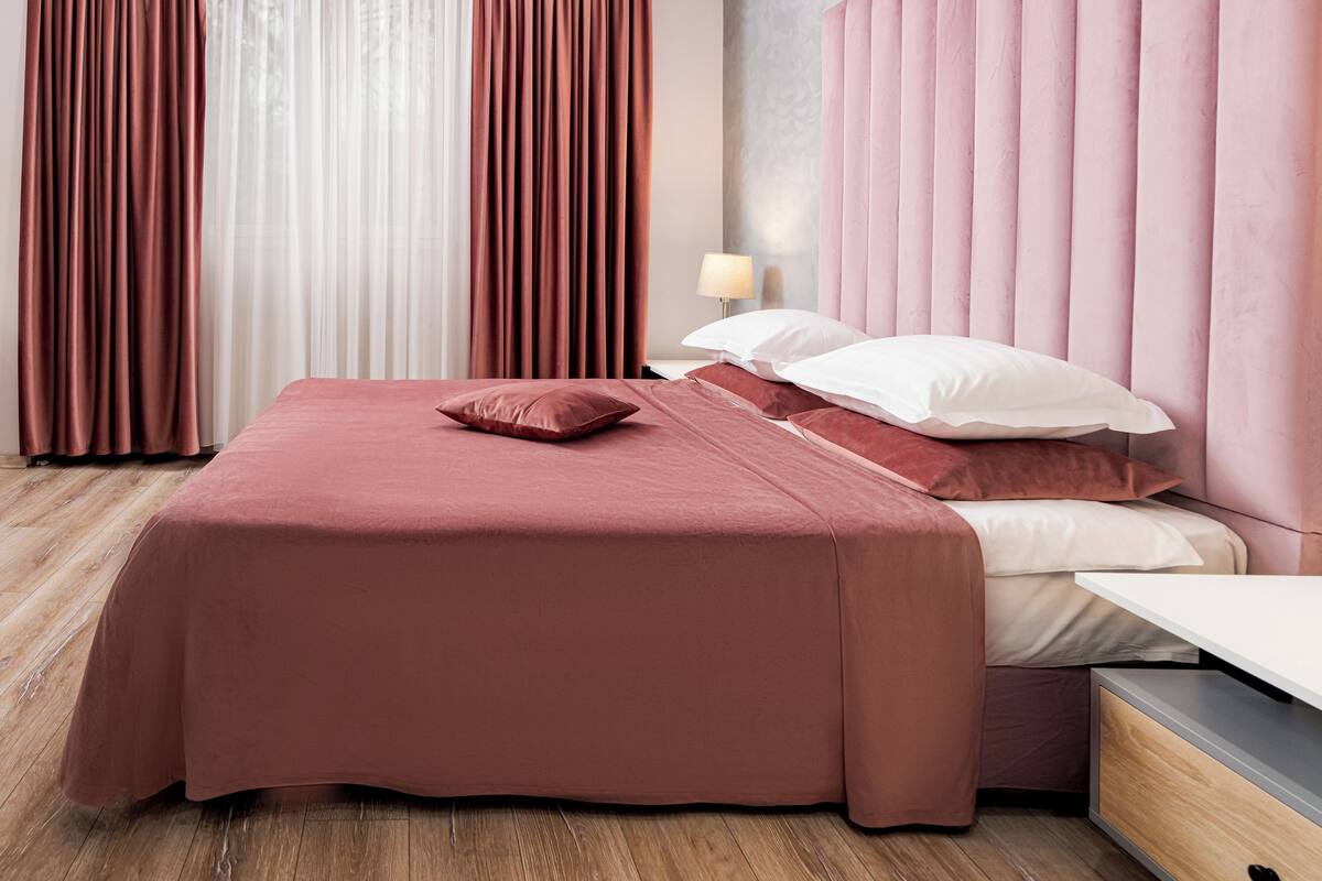 A neatly made bed with a pink bedspread and pillows, a bedside table with a lamp, and a wooden floor.