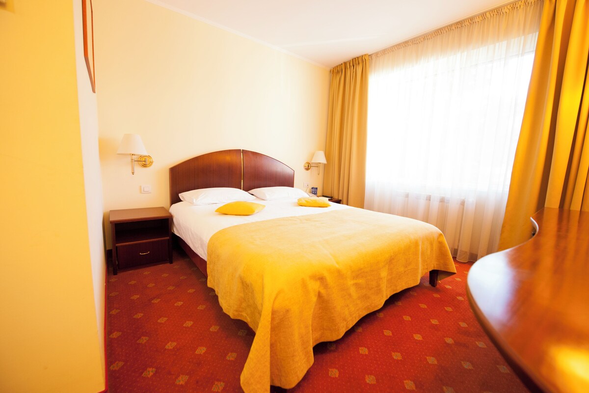 Ein Hotelzimmer mit Bett, Nachttisch, Lampe und Blick ins Freie durch ein Fenster mit Vorhängen.