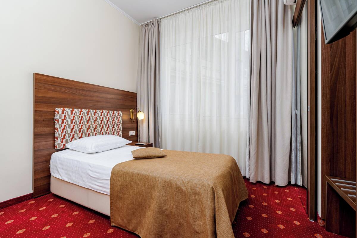 A hotel room with a bed, curtains, a TV, and a red carpeted floor.
