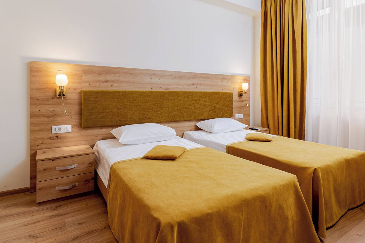 A hotel room with two beds, wooden headboard, bedside lamps, a nightstand, and a window with curtains.