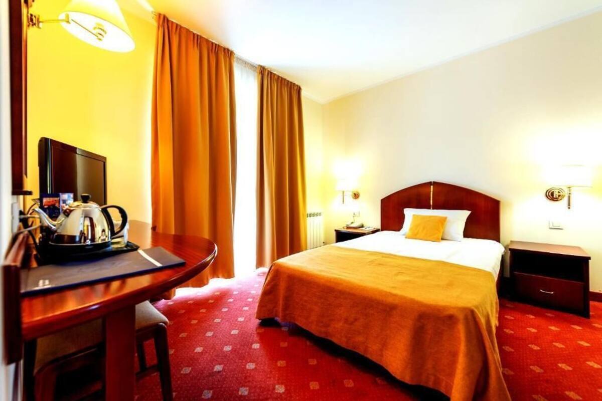 A hotel room with a bed, a desk with a kettle, a television, and two bedside lamps.