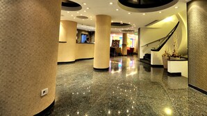 Un moderno vestíbulo de hotel con piso de mármol, columnas grandes, una recepción y una escalera.