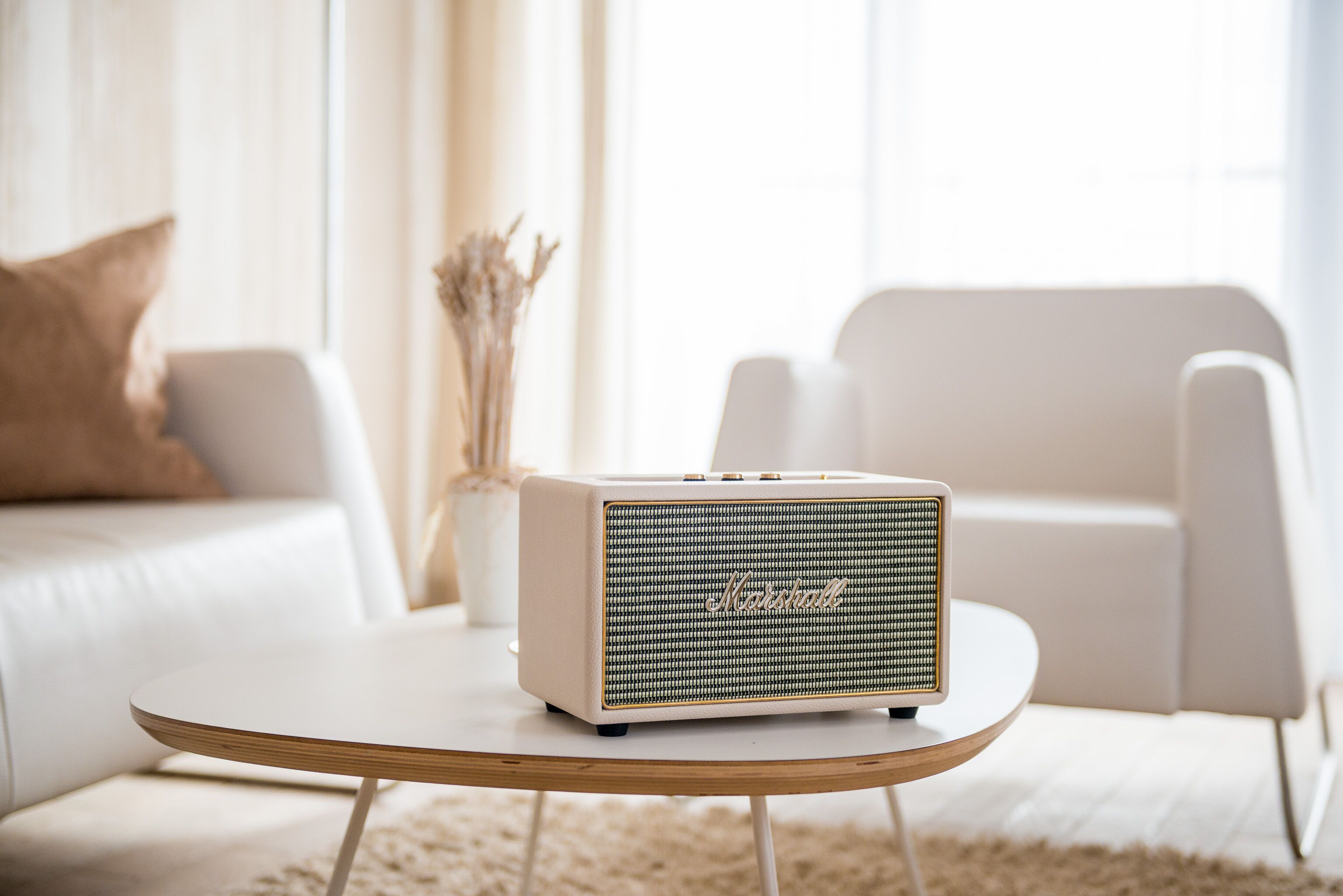 Een witte Marshall speaker op een ronde houten tafel in een moderne woonkamer met witte meubels en een vaas gedroogde bloemen.