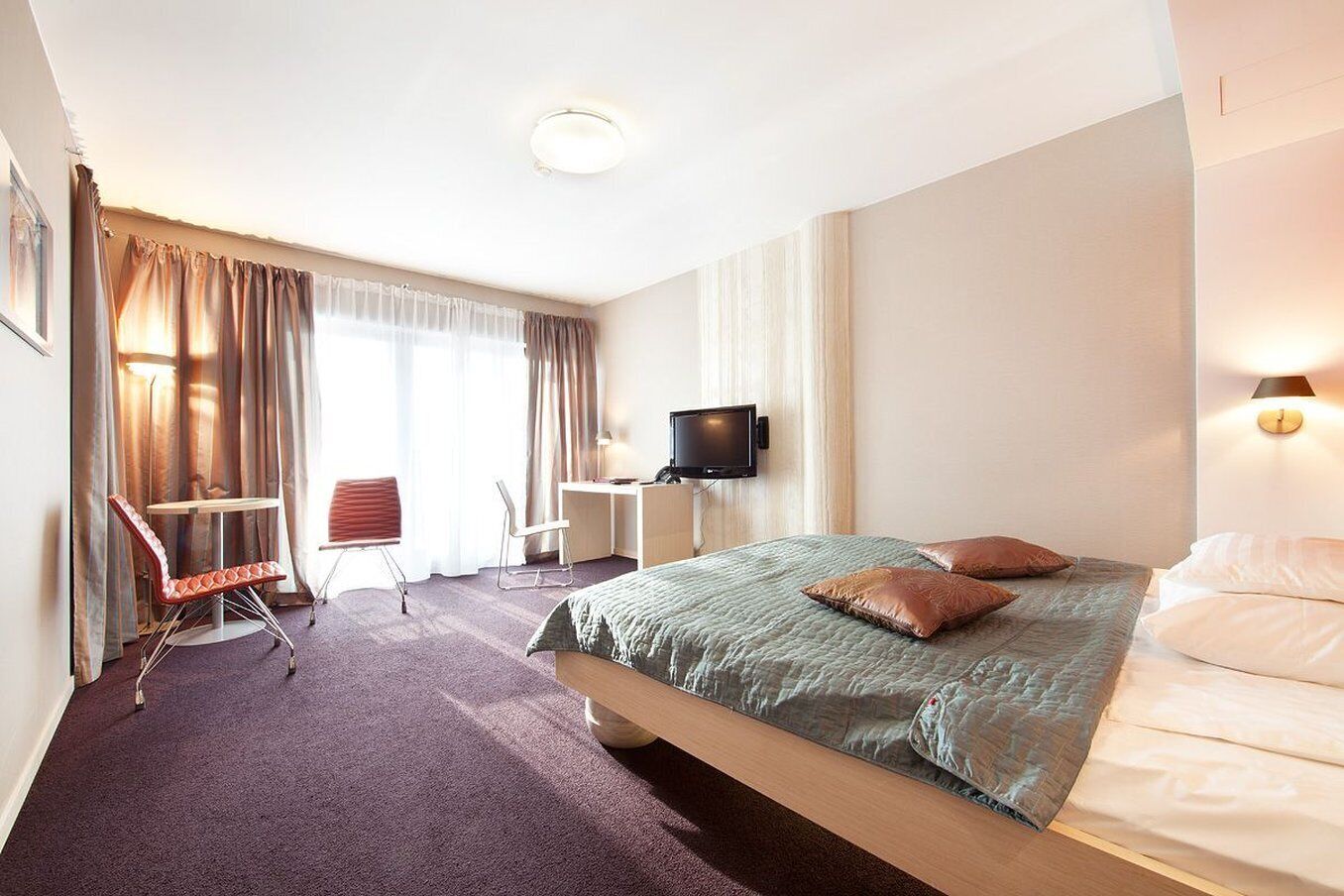 Habitación de hotel con cama, escritorio, silla, televisor y ventana con cortinas.