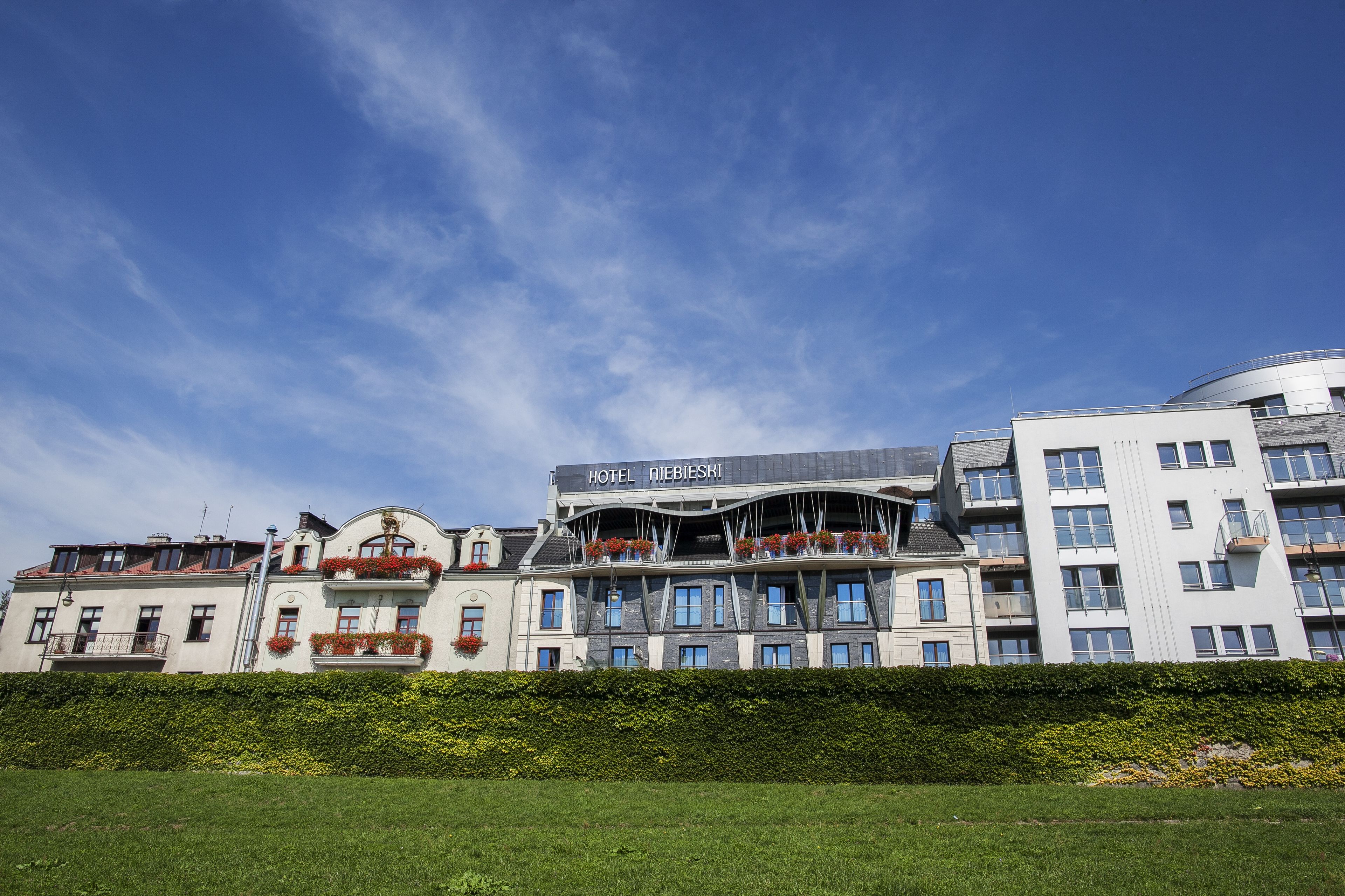 A hotel with a modern architectural design, featuring multiple balconies with flower boxes, a prominent sign, and a well-maintained garden in front.