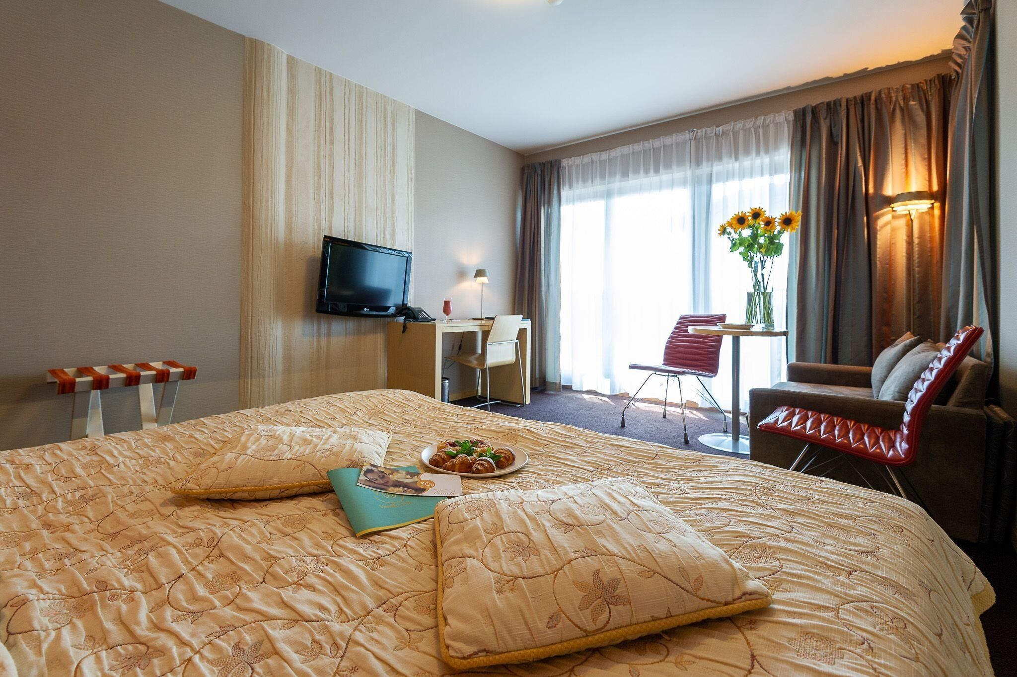 A hotel room with a large bed, a desk with a chair, a television, a dining table with a chair, and a window with curtains.