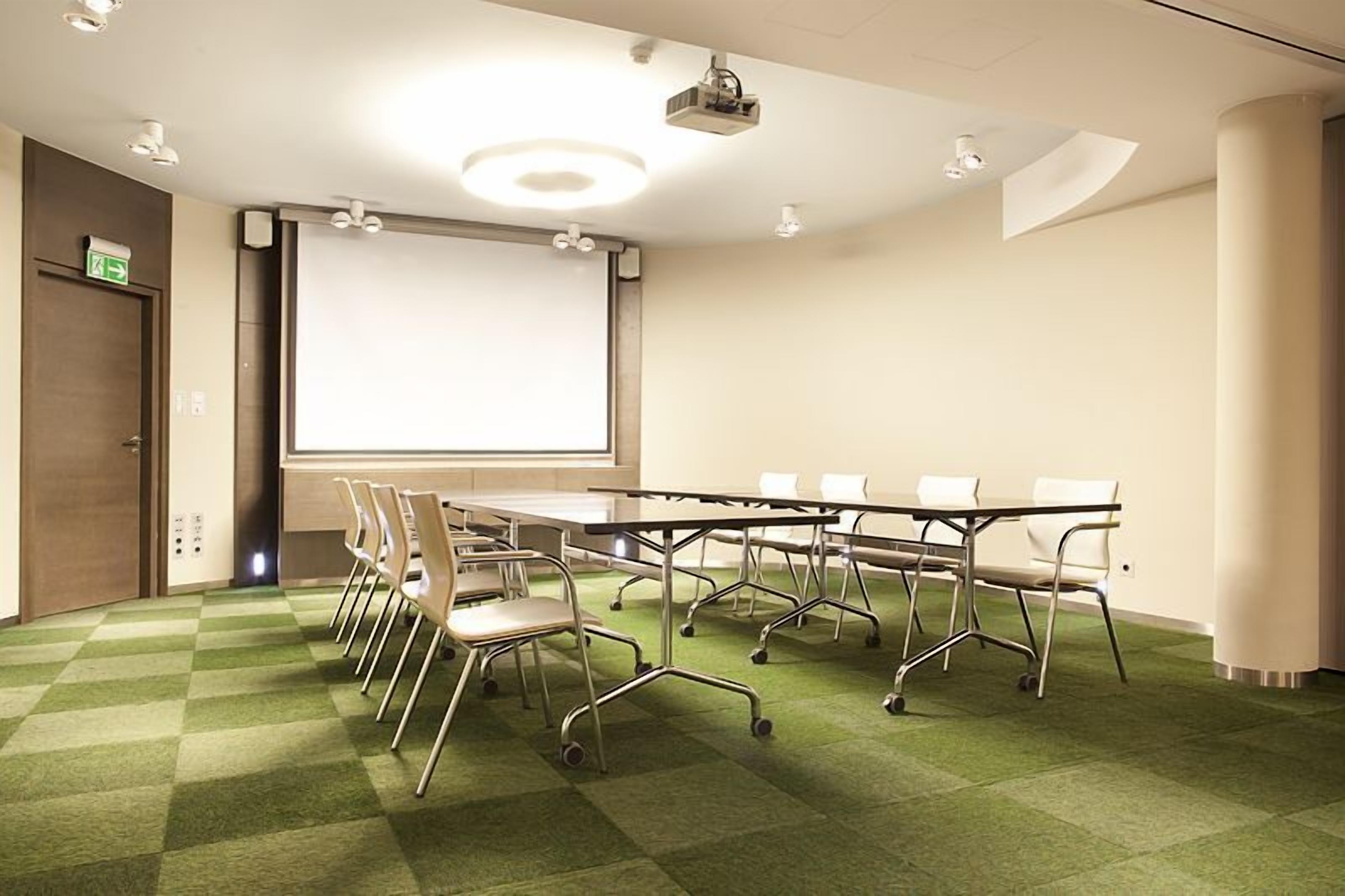 Een vergaderzaal met een grote tafel, stoelen, een scherm voor een beamer en een vloerbedekking.