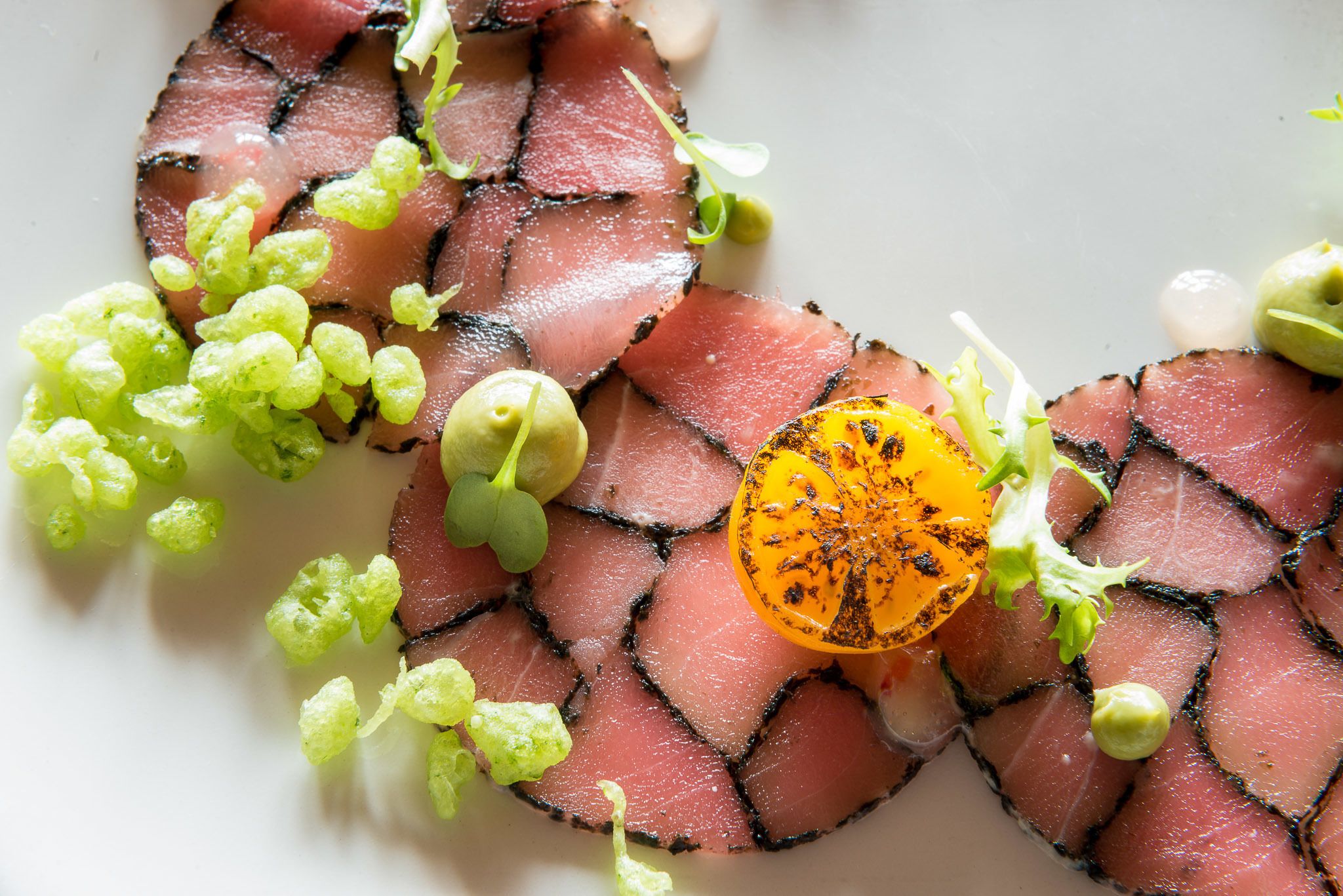 Um prato com fatias finas de carne dispostas em forma de círculo, decorado com folhas verdes e uma fatia de fruta grelhada.