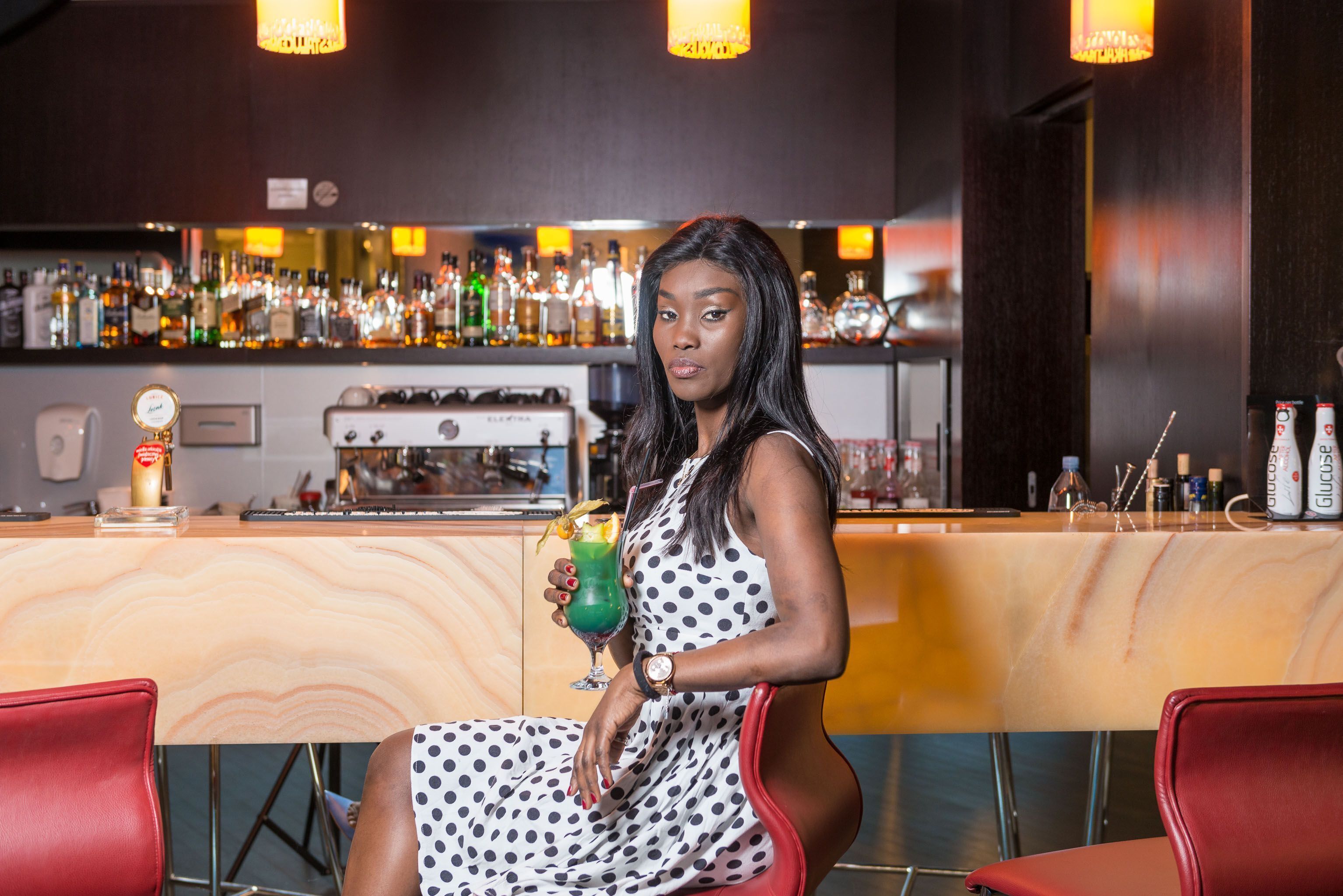 A woman seated at a bar with a drink in hand, a bar stocked with various bottles, and a cash register.