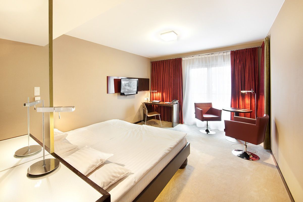 A hotel room with a bed, desk, chair, TV, and a bedside table with a lamp.