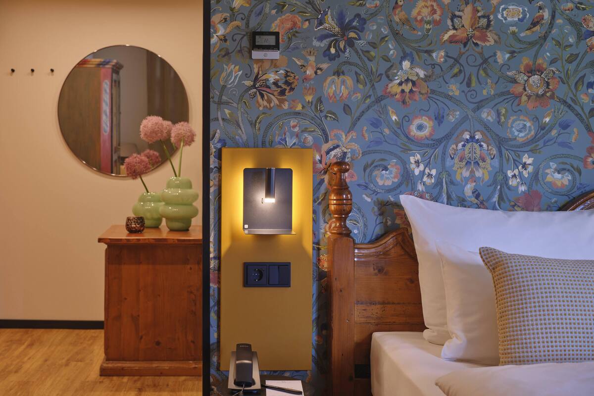A hotel room with a wooden bed, a bedside table with a phone, a mirror, and a floral wallpaper.