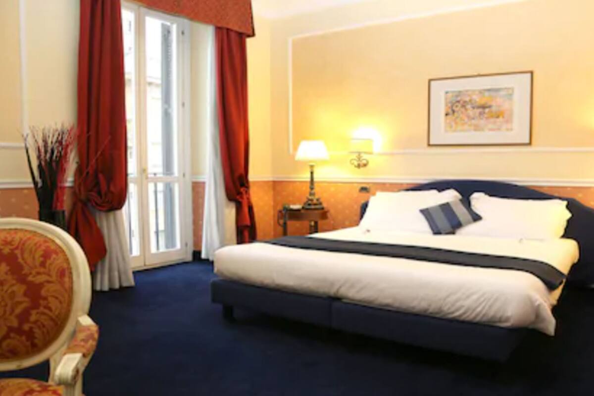 A hotel room with a large bed, two bedside lamps, a painting on the wall, and a chair with a floral pattern.