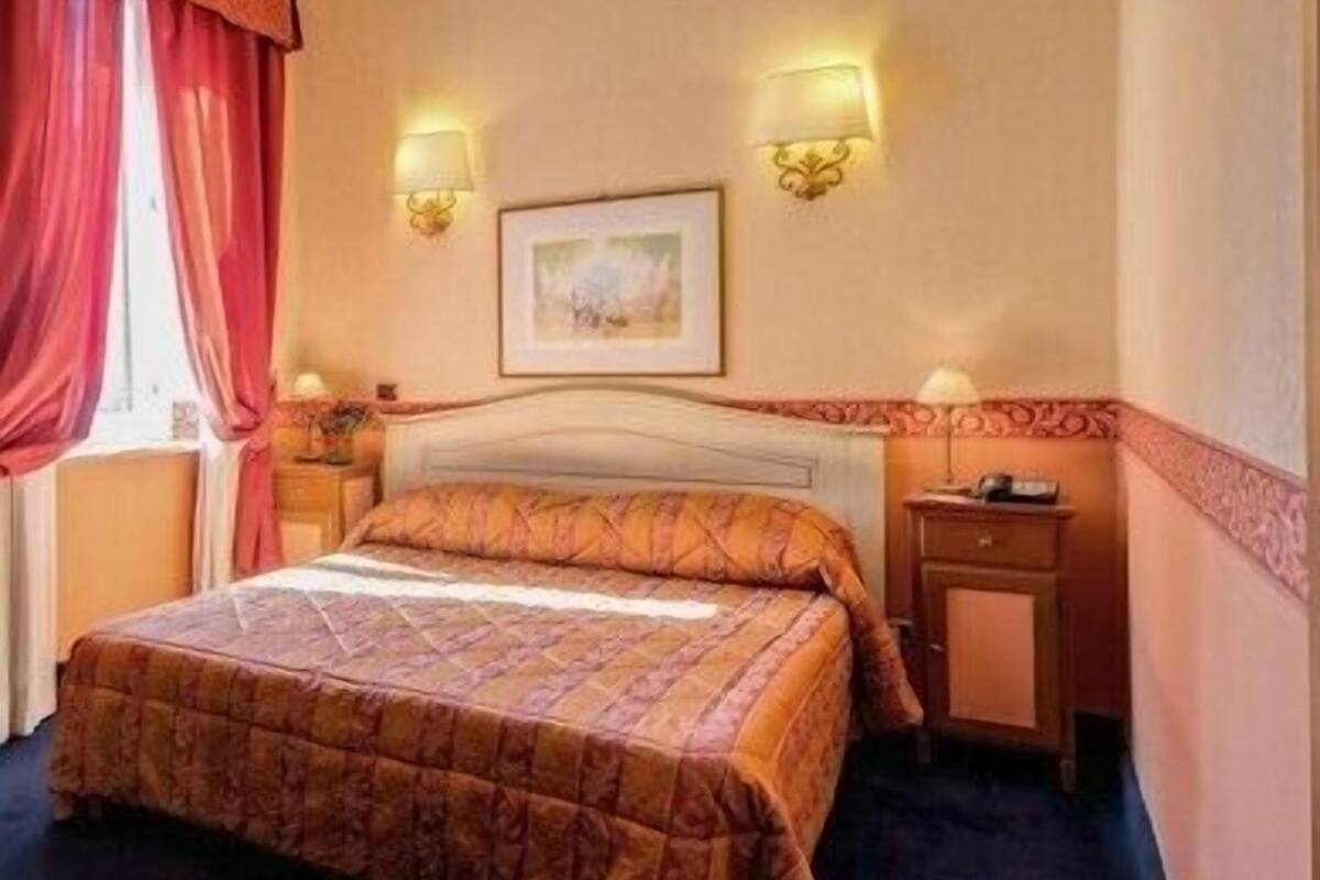 A hotel room with a bed, two bedside lamps, a nightstand, a framed picture on the wall, and a window with red curtains.