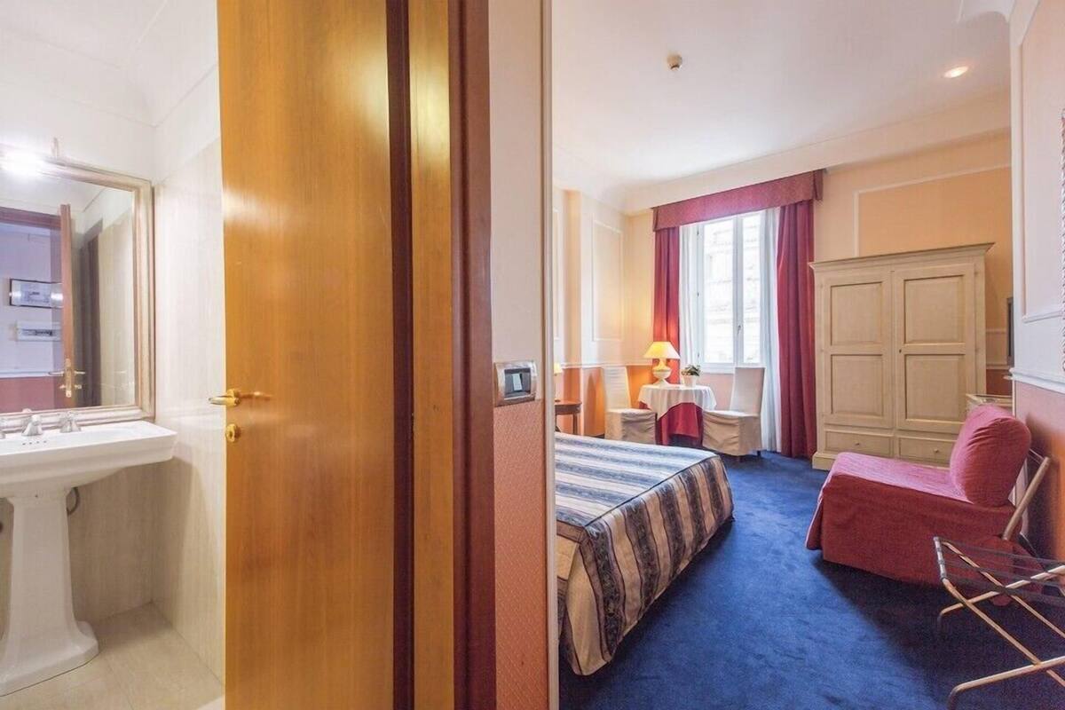 A hotel room with a bed, a desk, a chair, a wardrobe, and a bathroom with a sink and mirror.
