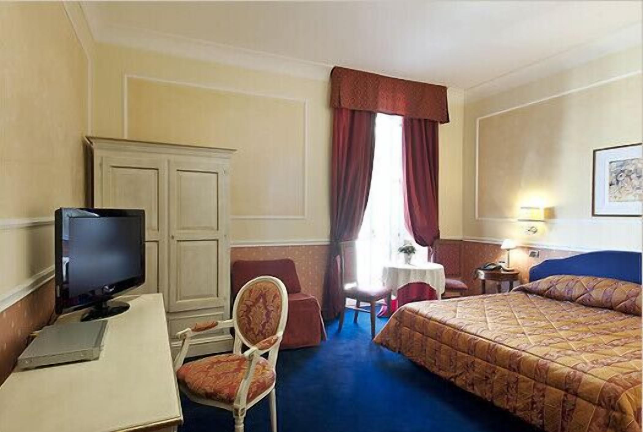 Habitación de hotel con cama, escritorio con televisor, silla, armario y ventana con cortinas.