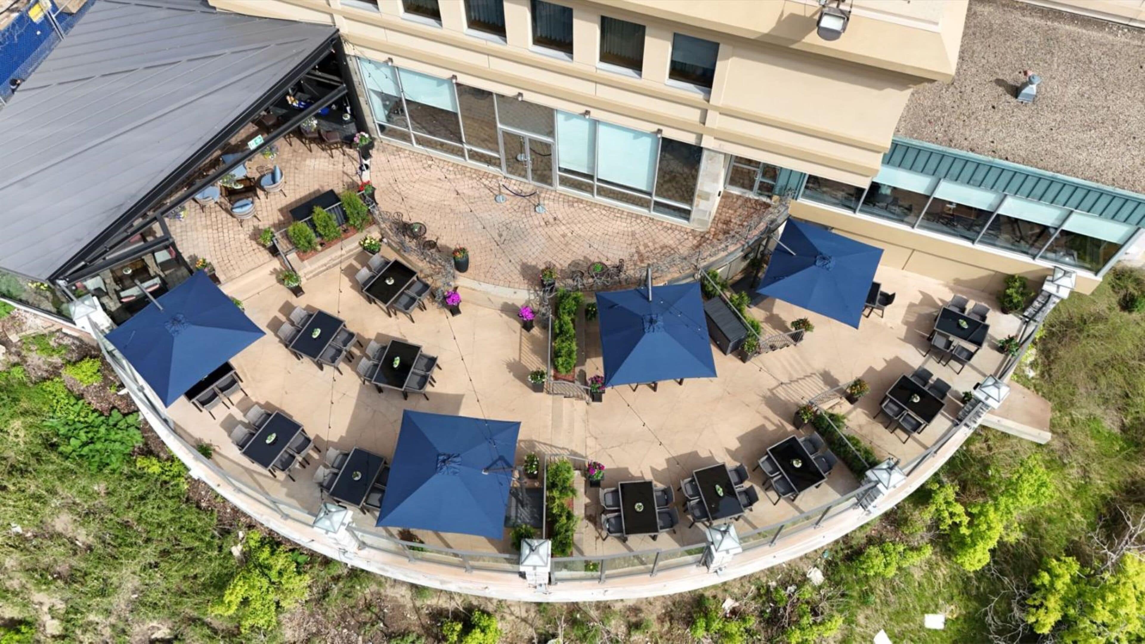 Un espace extérieur aménagé en terrasse, avec plusieurs tables et des parasols bleus, le tout entouré d’une balustrade en verre incurvée.