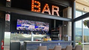 A bar with a large screen displaying sports events, a counter with bottles, and bar stools. - Courtyard by Marriott Edmonton Downtown (Edmonton)
