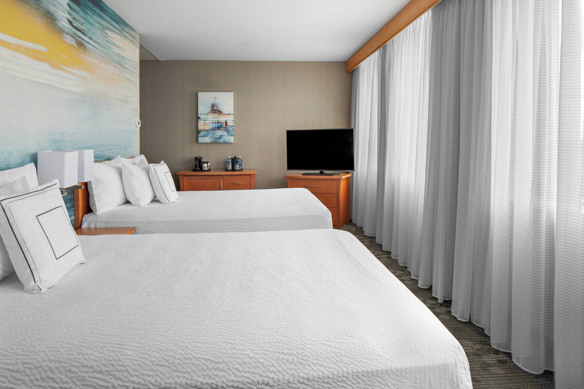 A hotel room with two beds, a flat-screen TV, a painting on the wall, and a large window with curtains.
