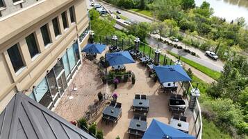 An outdoor patio area with multiple tables and umbrellas, surrounded by greenery and a view of a road and vehicles.