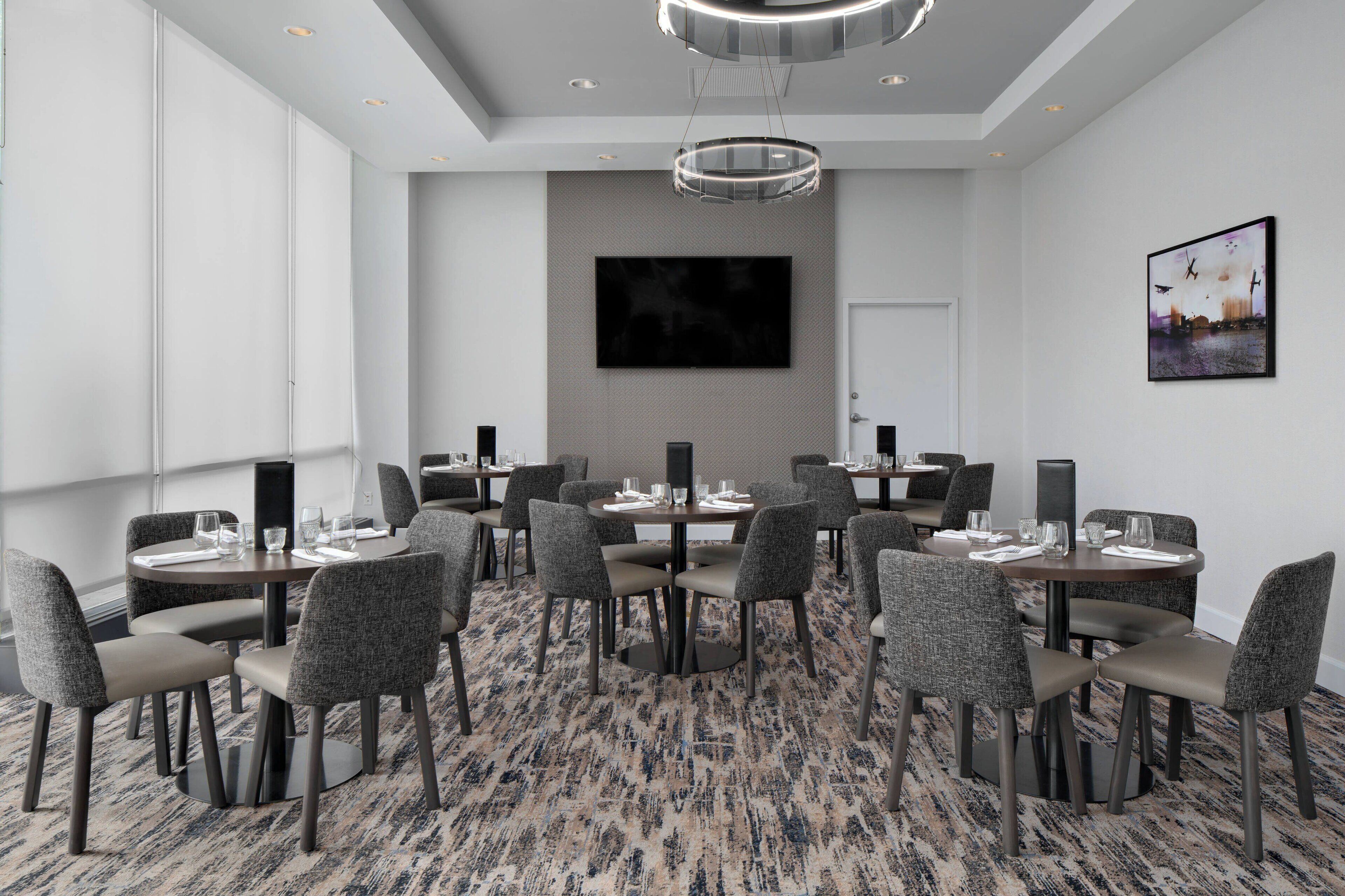 A conference room with round tables and grey upholstered chairs, a large flat-screen TV on the wall, and a patterned carpet.