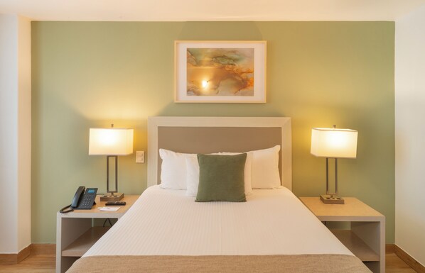 A hotel room with a bed, two bedside lamps, a telephone, and a painting on the wall.
