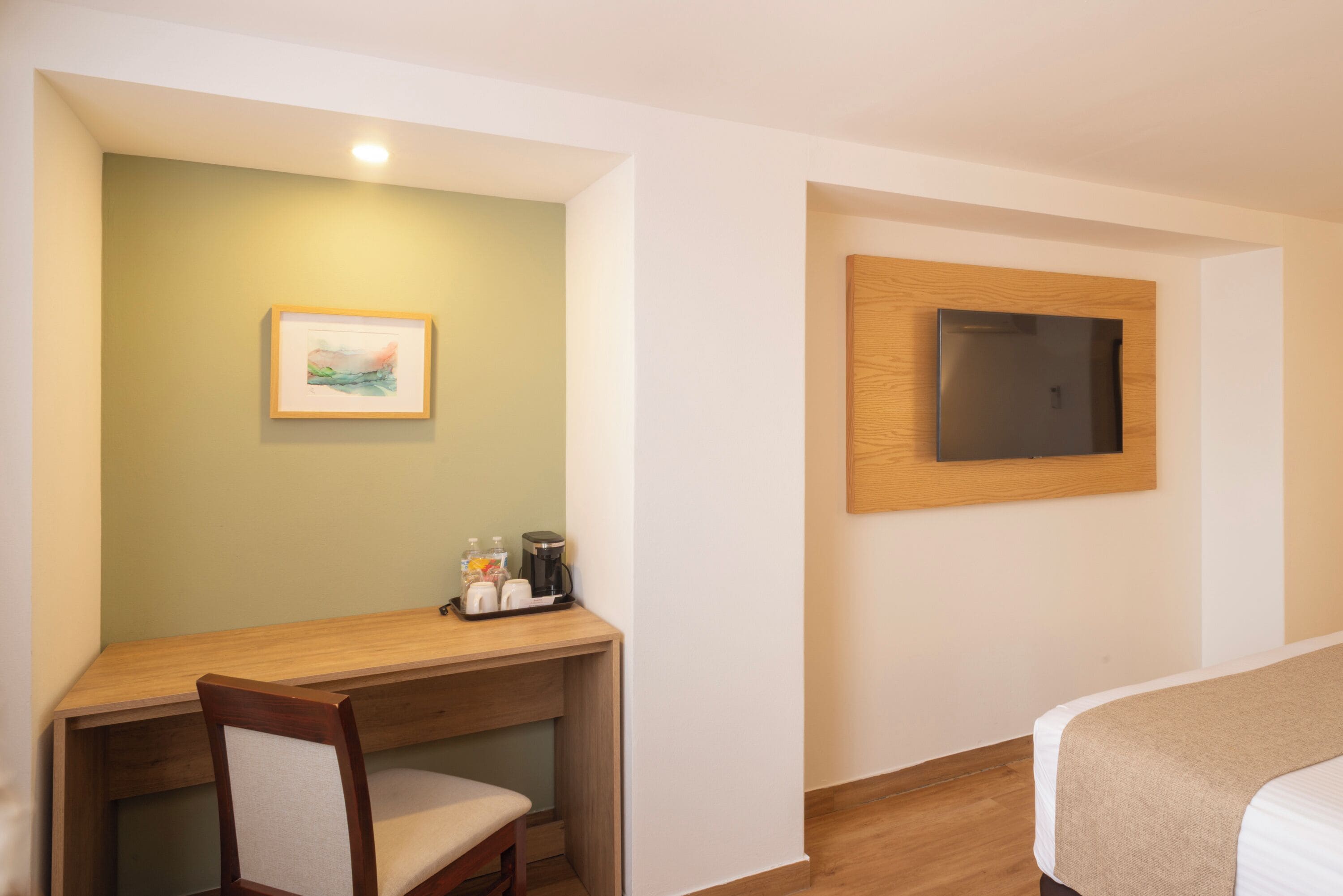 Habitación de hotel con escritorio de madera, una silla, una cama, televisor montado en la pared y un cuadro enmarcado.