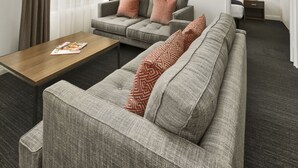 A modern hotel room with a grey sofa set, a wooden coffee table, a framed picture on the wall, and a door leading to a bathroom. - Quest Dandenong (Dandenong)