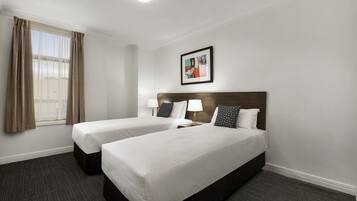 A hotel room with two single beds, a window with curtains, a framed artwork on the wall, and a carpeted floor.