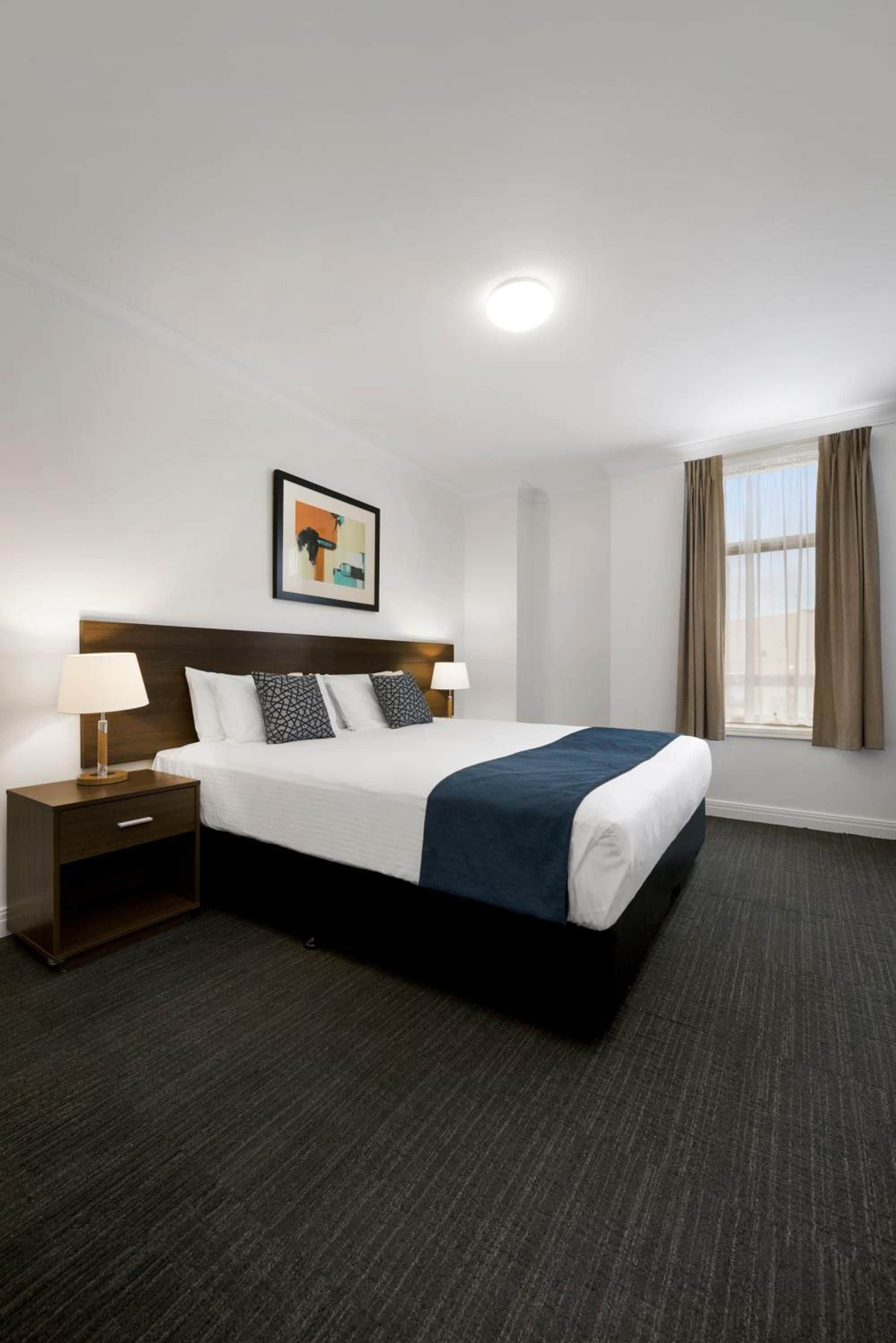 Habitación de hotel con una cama grande, una mesita de noche, una lámpara, un cuadro en la pared y una ventana con cortinas.