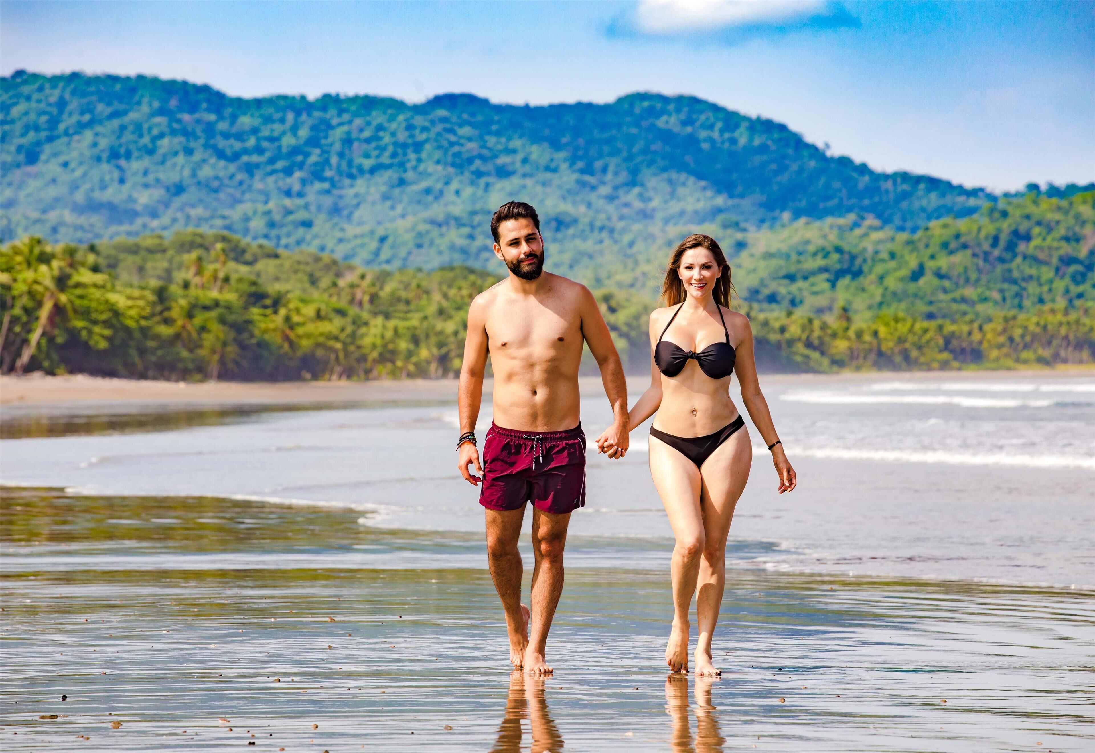 A couple walking on a beach with a forested hill in the background.