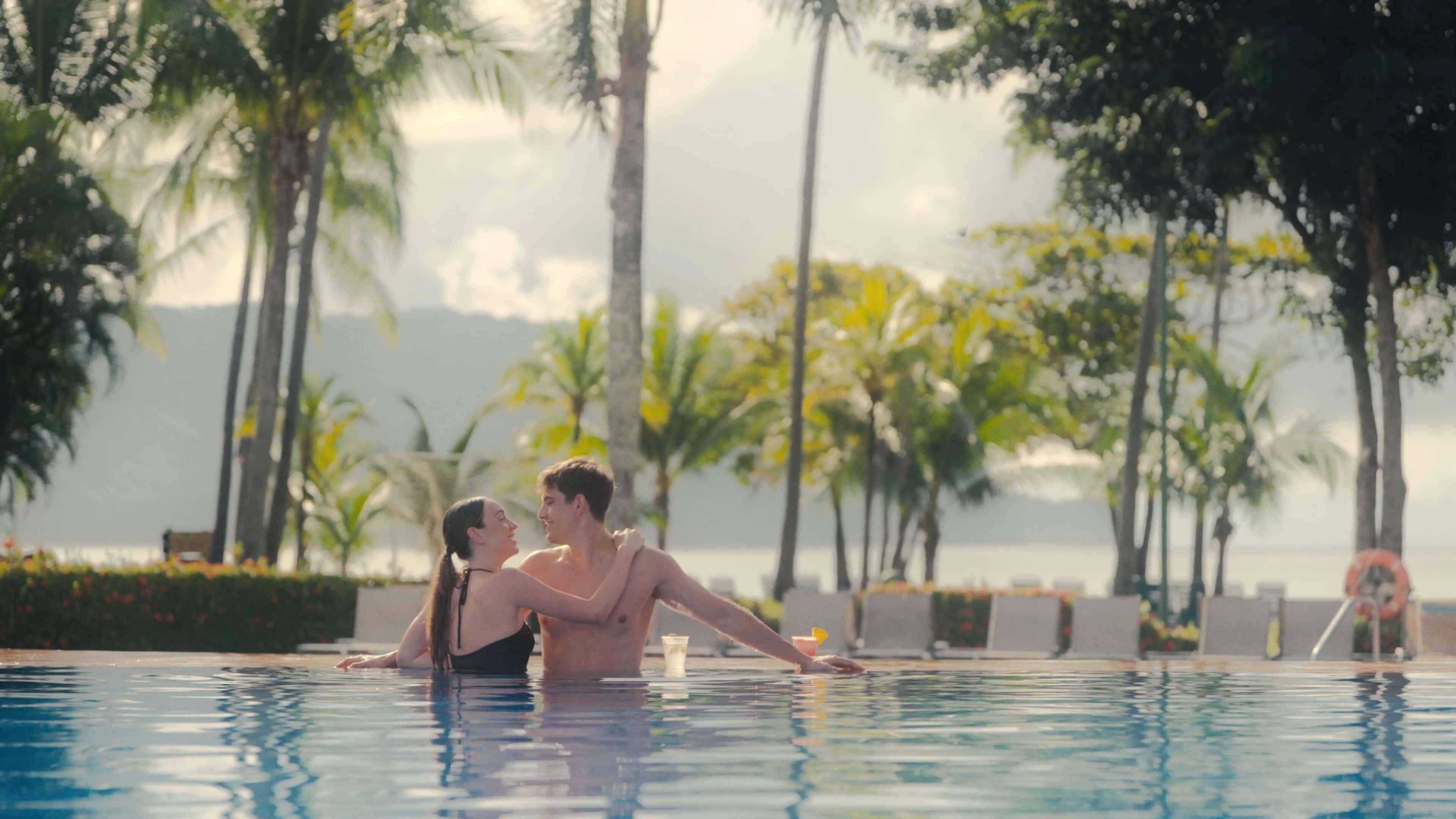A couple in a swimming pool with palm trees and a view of the sea in the background.