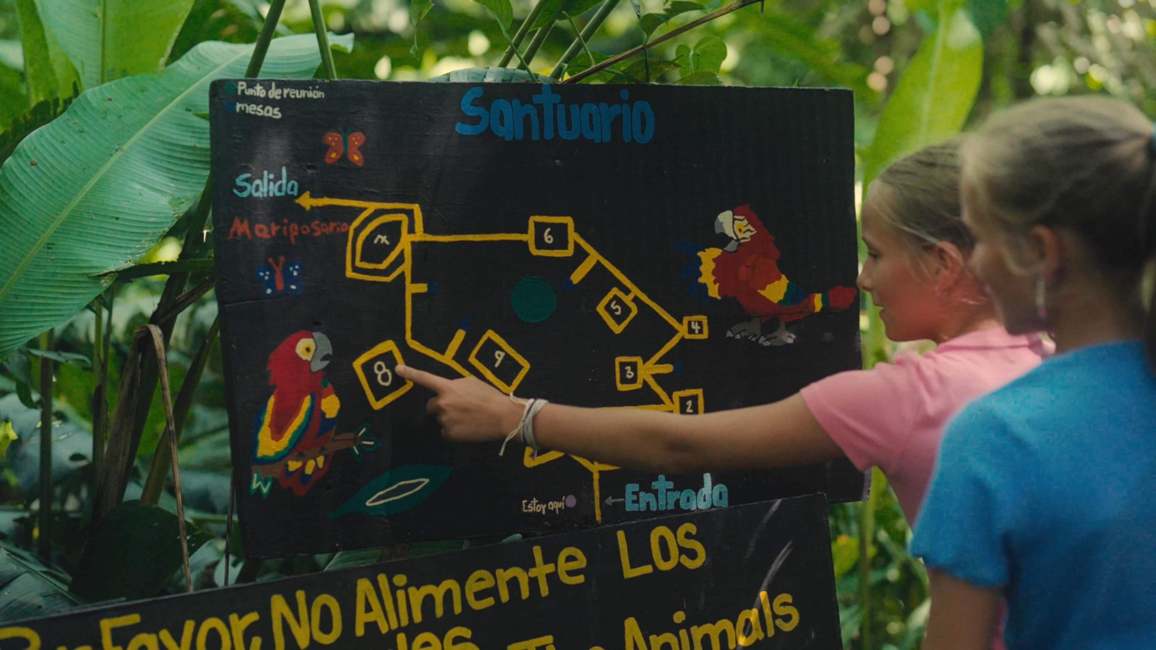 A person pointing at a black board with a map and text, surrounded by greenery.