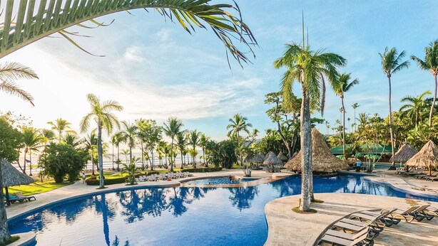 A resort pool with lounge chairs, palm trees, and thatched-roof huts. - Barceló Tambor (Paquera)