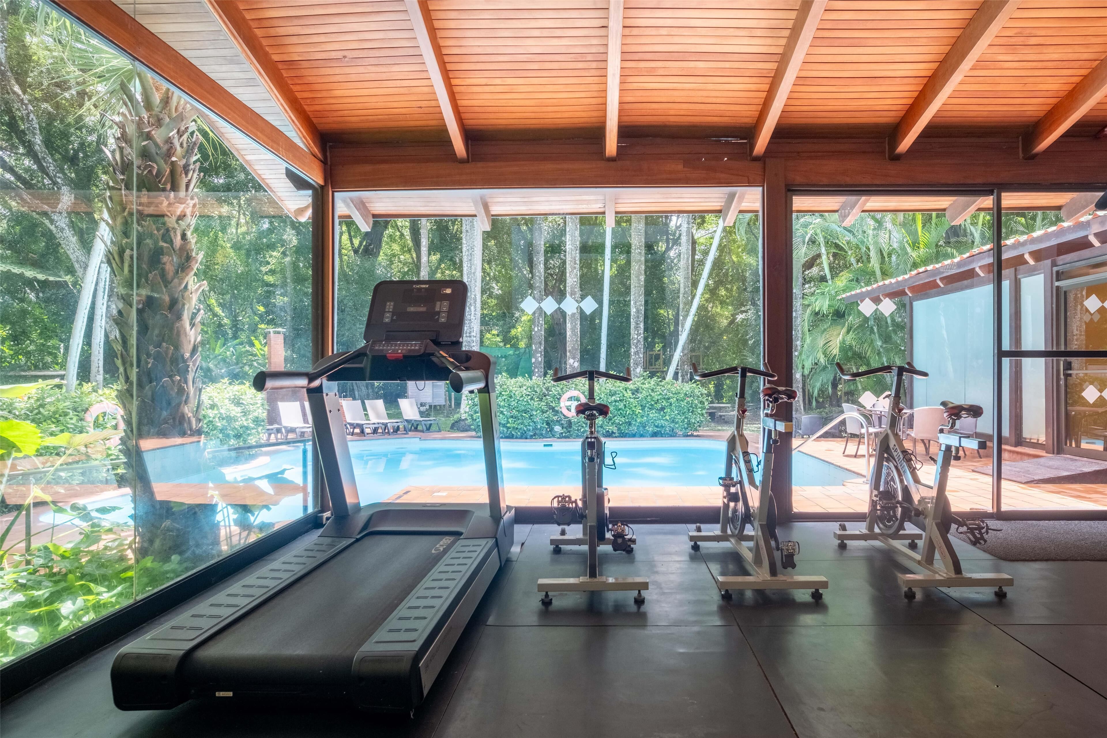 Un gimnasio con caminadora, bicicletas estáticas y máquina de remo, con vistas a una piscina y abundante vegetación.