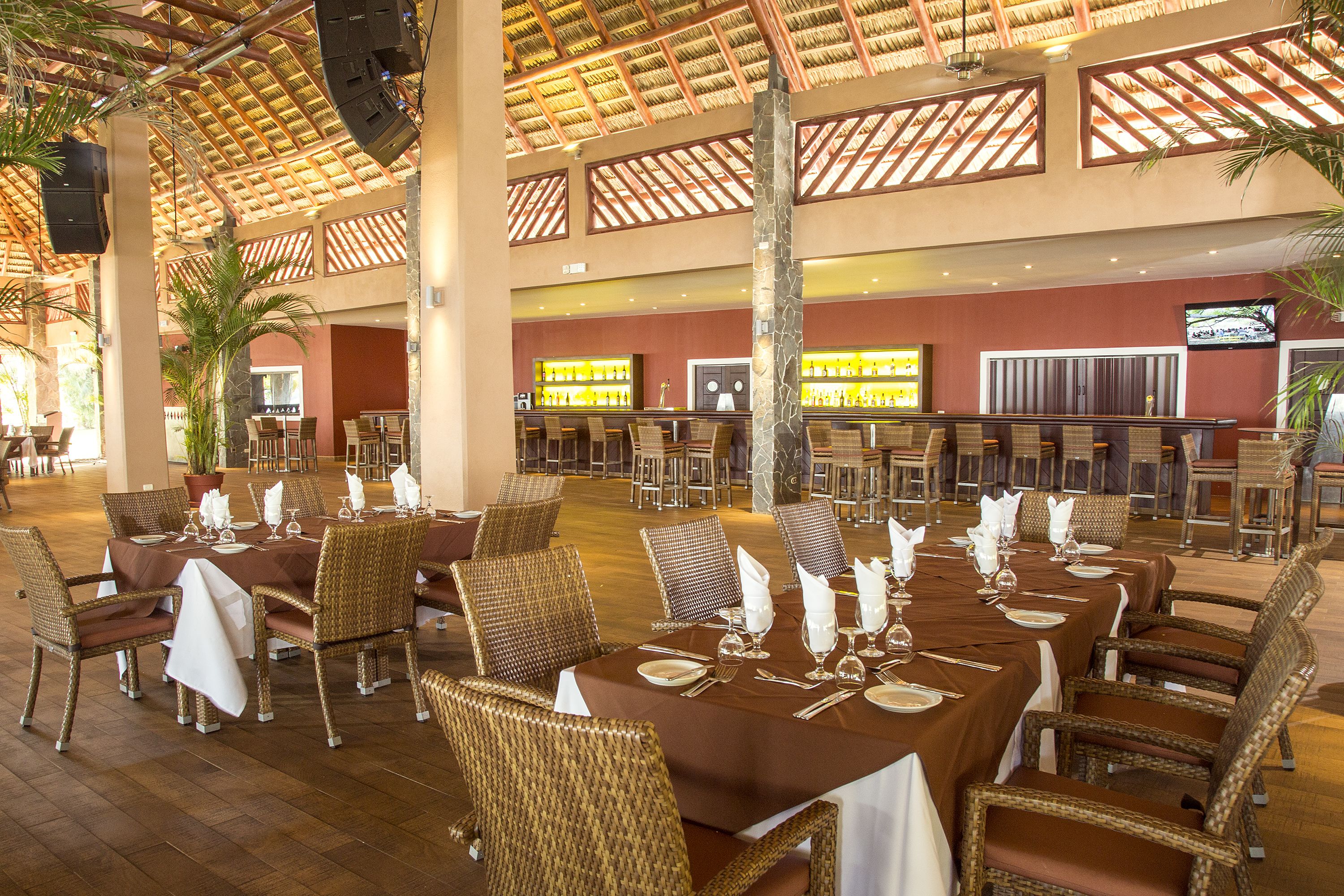 Amplia zona de comedor interior con varias mesas dispuestas para recibir a los huéspedes, con sillas de mimbre y un techo alto con diseño de techo de paja.