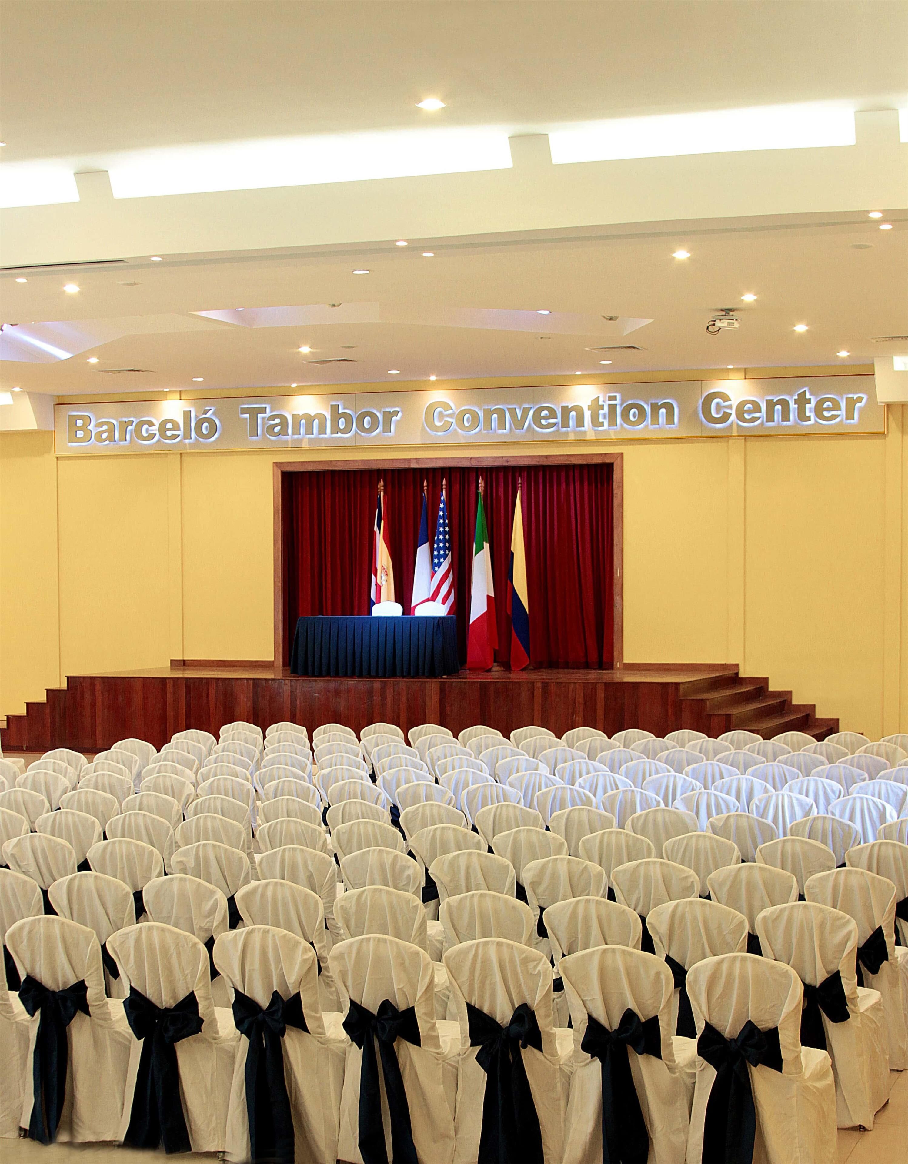 Ett konferensrum med rader av stolar, en scen med flaggor och en skylt med texten ”Barceló Tambor Convention Center”.