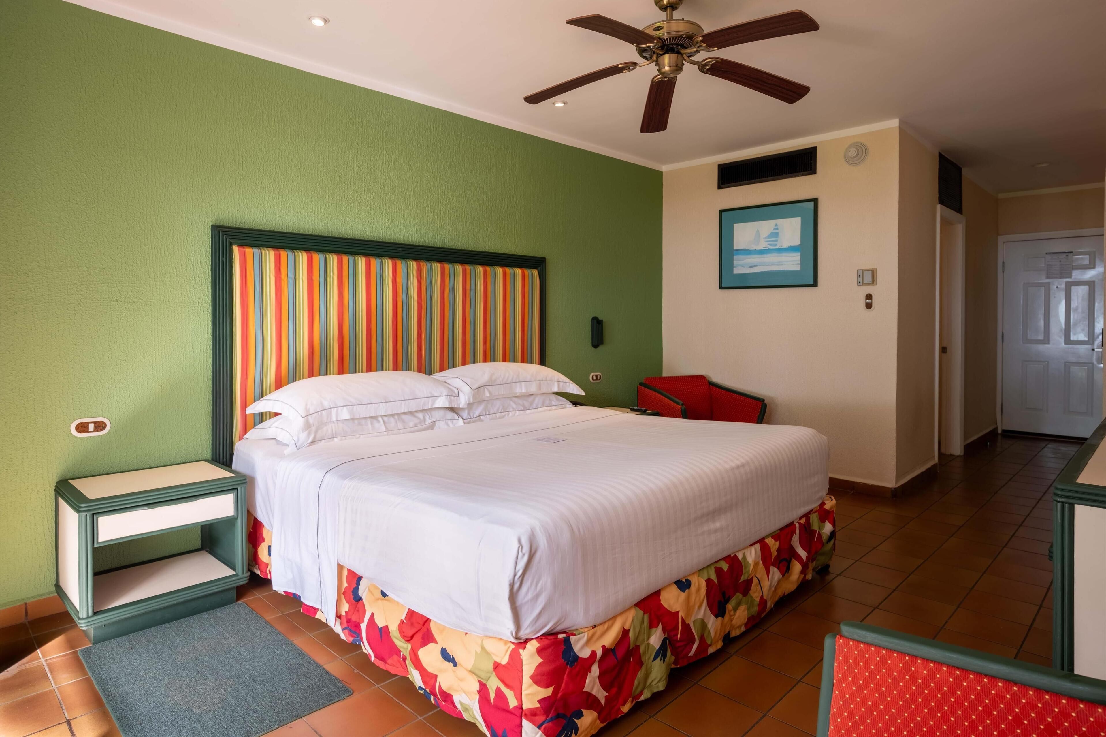 Une chambre d’hôtel avec un lit, une table de chevet, une chaise rouge et un ventilateur de plafond.