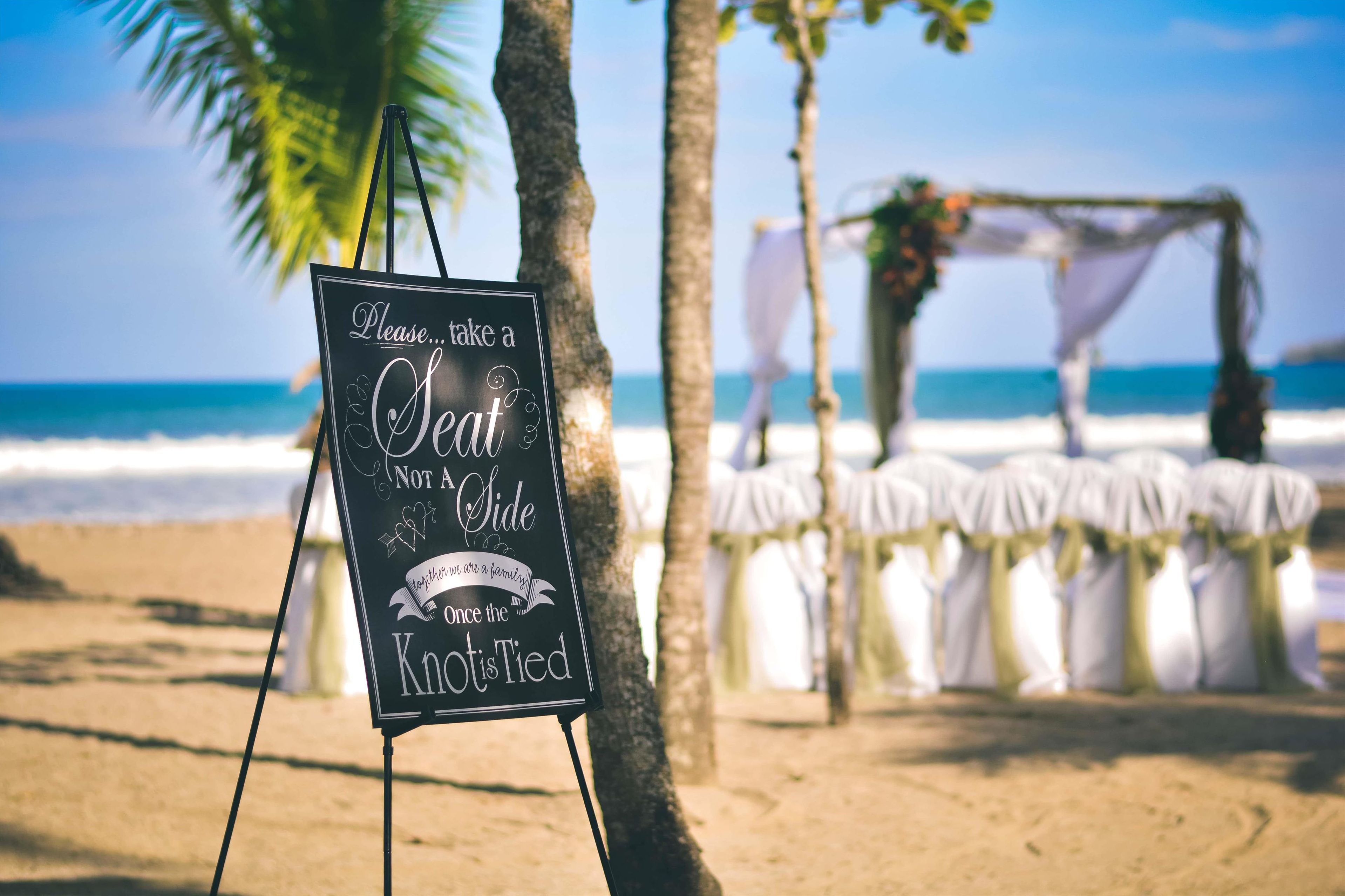 A beach wedding setup with a sign that reads "Please... take a Seat Not A Side Once the Knot is Tied".